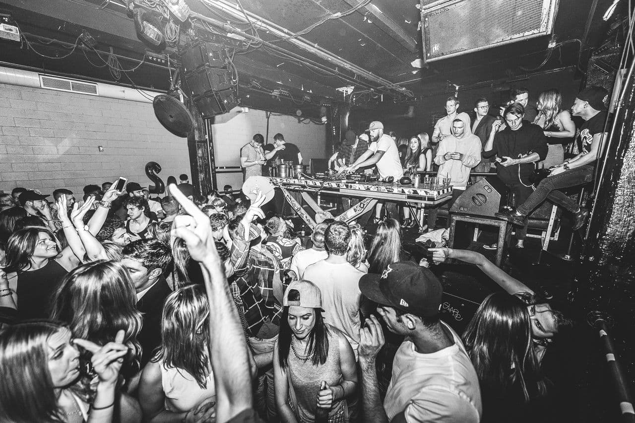 A packed warehouse party crowd in front of a DJ booth